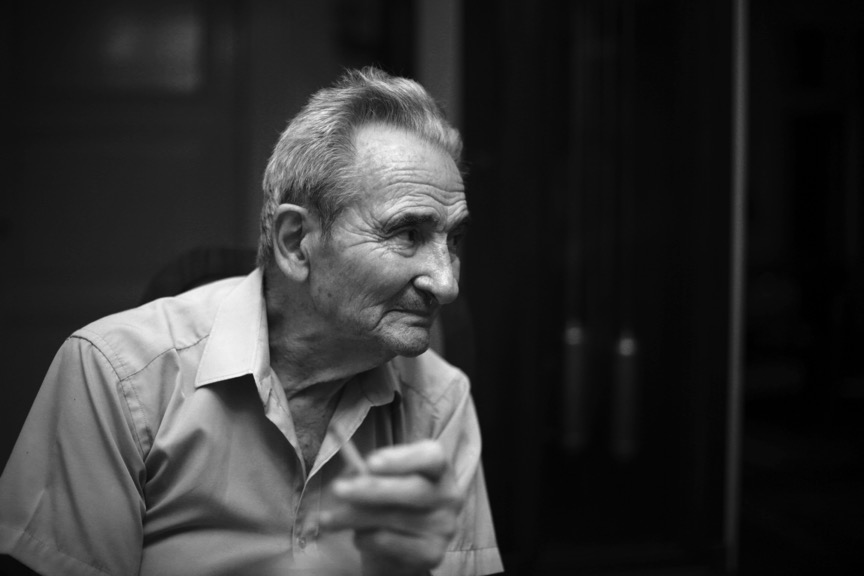 Photographie en noir et blanc de Licu regardant vers la droite de l’image. Il tient une cigarette à la main. En fond, légèrement flou, le balancier d’une pendule.